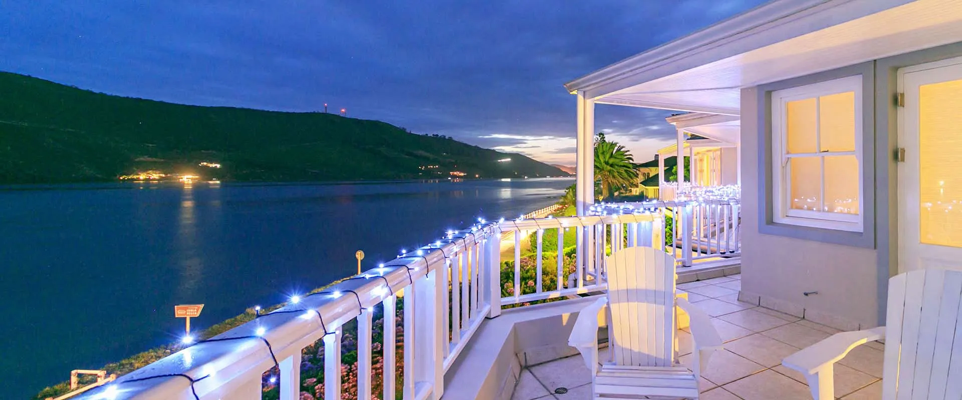 White chairs on a balcony overlooking a lake at dusk with lights on railing
