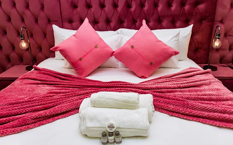 Bed with pink pillows and blanket white towels and toiletries on a red headboard