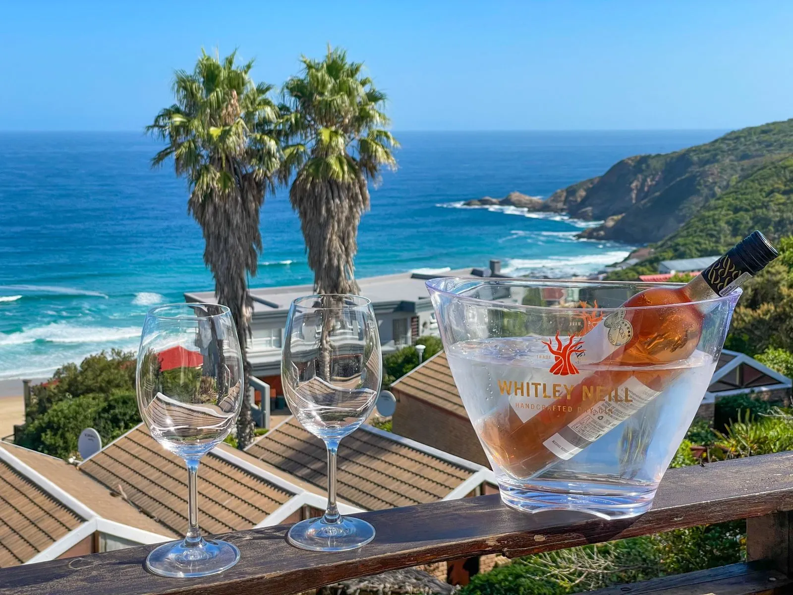 Two wine glasses and a bucket with a bottle overlooking ocean and palm trees