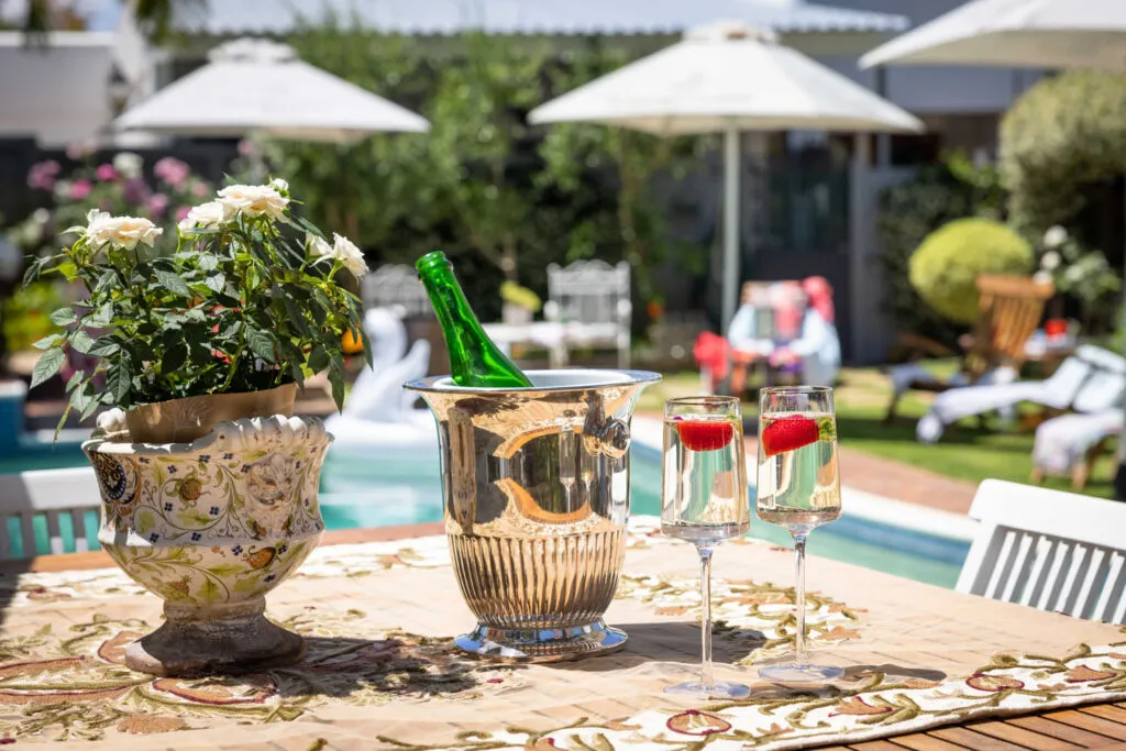 Champagne in ice bucket and glasses on table with pool and garden in background