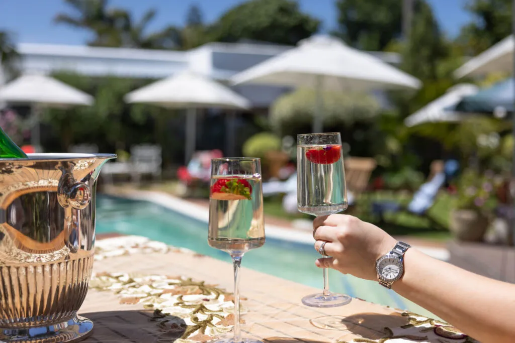 Two champagne glasses with strawberries by a pool with umbrellas in the background
