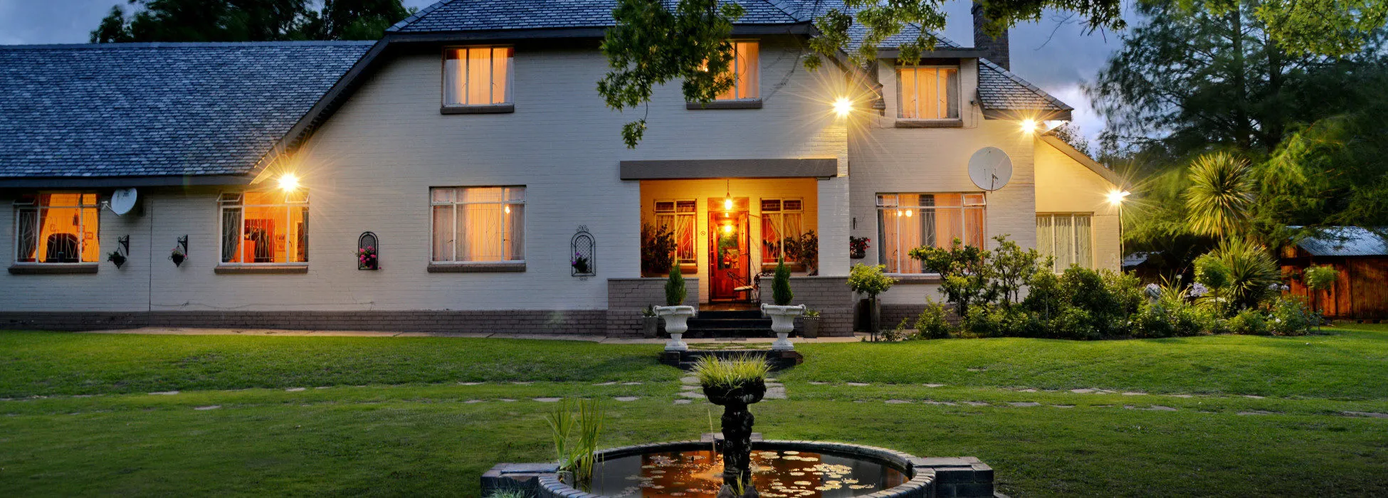 Large house with illuminated windows and a fountain in the front yard at night