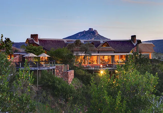 Luxury lodge with thatched roofs and mountain backdrop at dusk