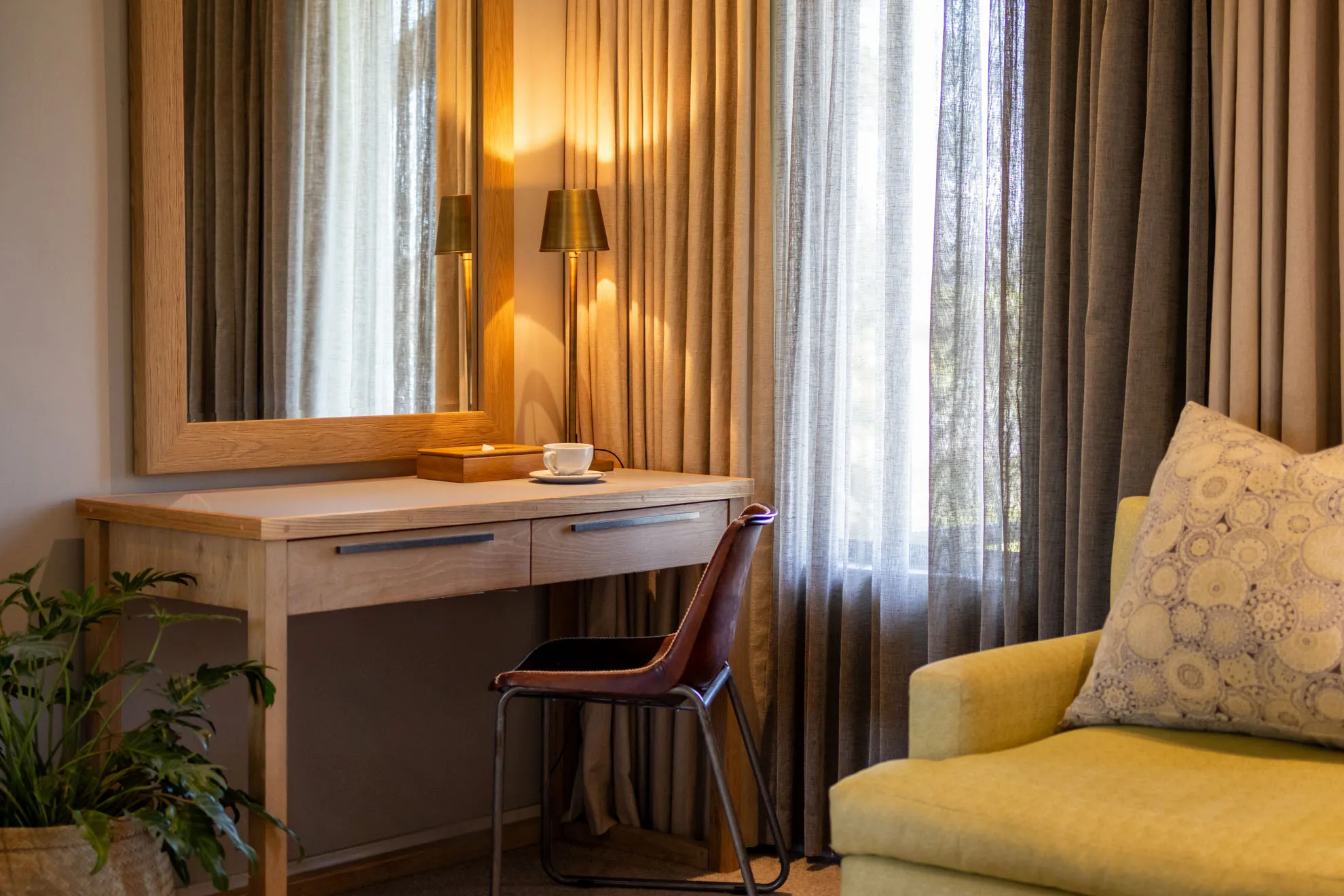 Wooden desk with chair lamp and cup beside yellow sofa with patterned pillow