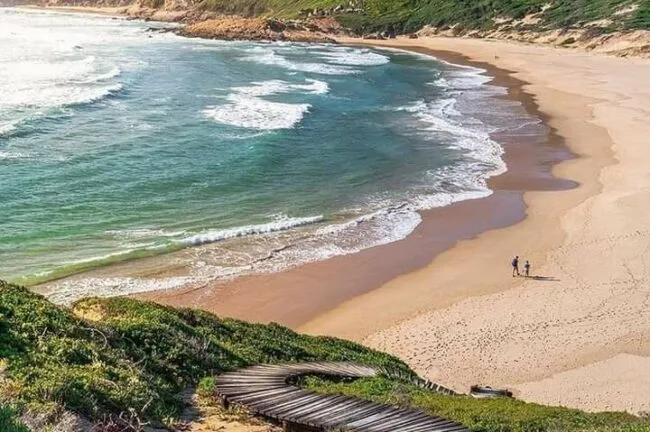 A sandy beach with waves two people walking and a wooden pathway