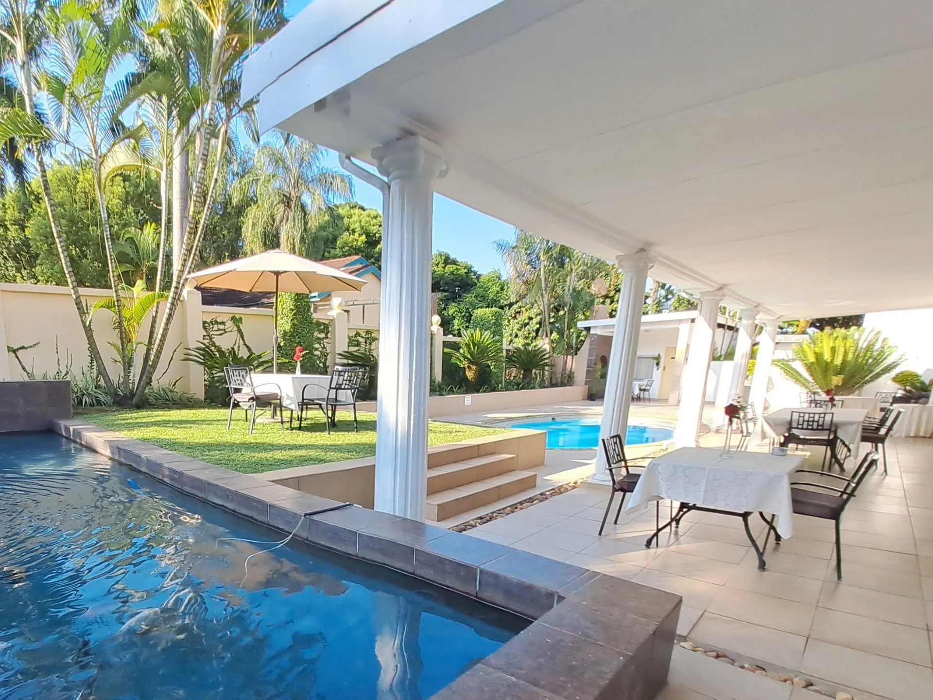 Outdoor dining area with tables chairs and a swimming pool surrounded by greenery