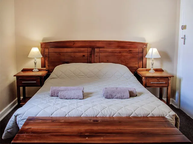 Wooden bed with white quilt two bedside tables with lamps and a wooden bench