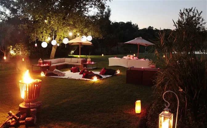 Outdoor evening setup with lanterns cushions and tables under trees near water