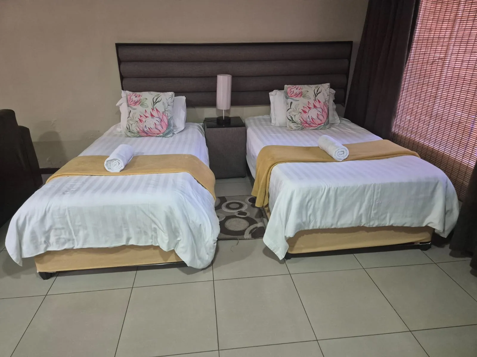 Two neatly made single beds in a bedroom with tiled floor and window blinds