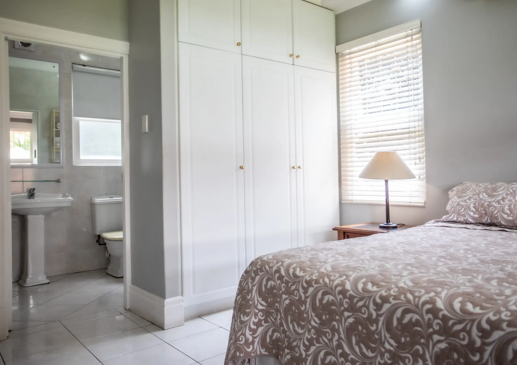 Bedroom with patterned bedspread wardrobe nightstand with lamp and adjacent bathroom