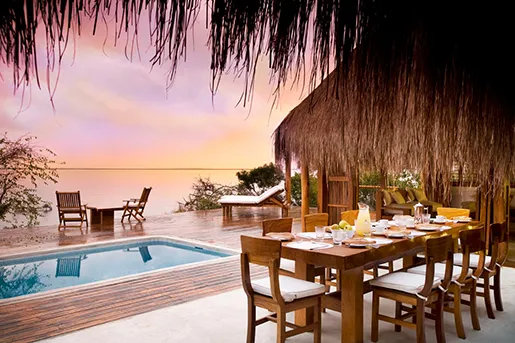 Wooden dining area with pool and sunset view under a thatched roof