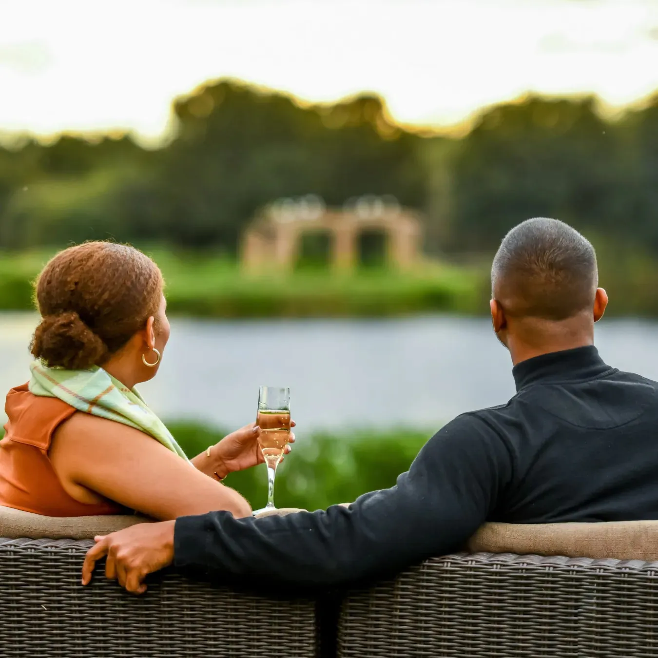 Couple sitting on a wicker sofa by a lake one holding a wine glass