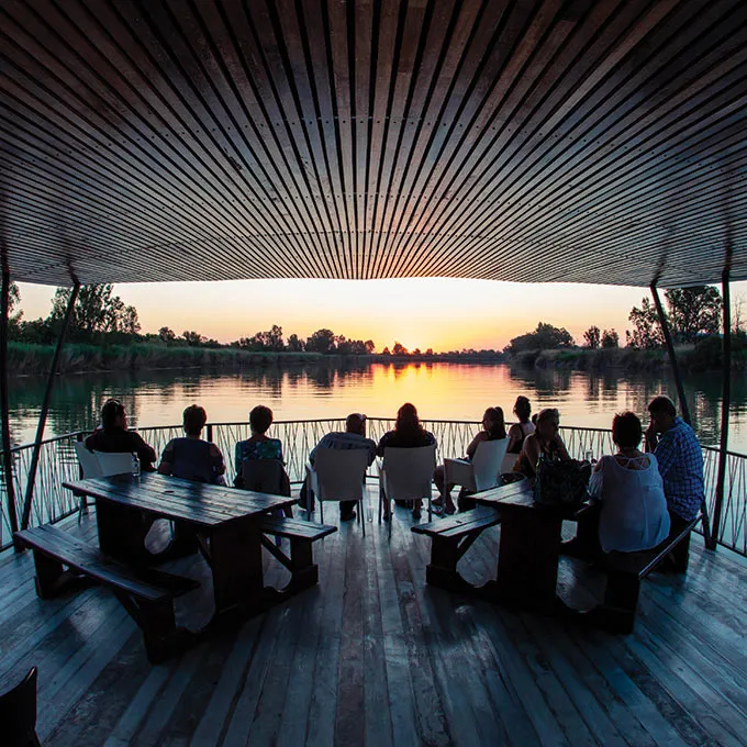 People sitting at tables on a wooden deck at sunset by a lake