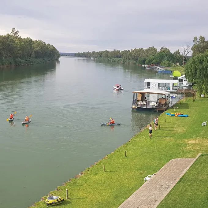 People kayaking on a river near a houseboat and grassy area