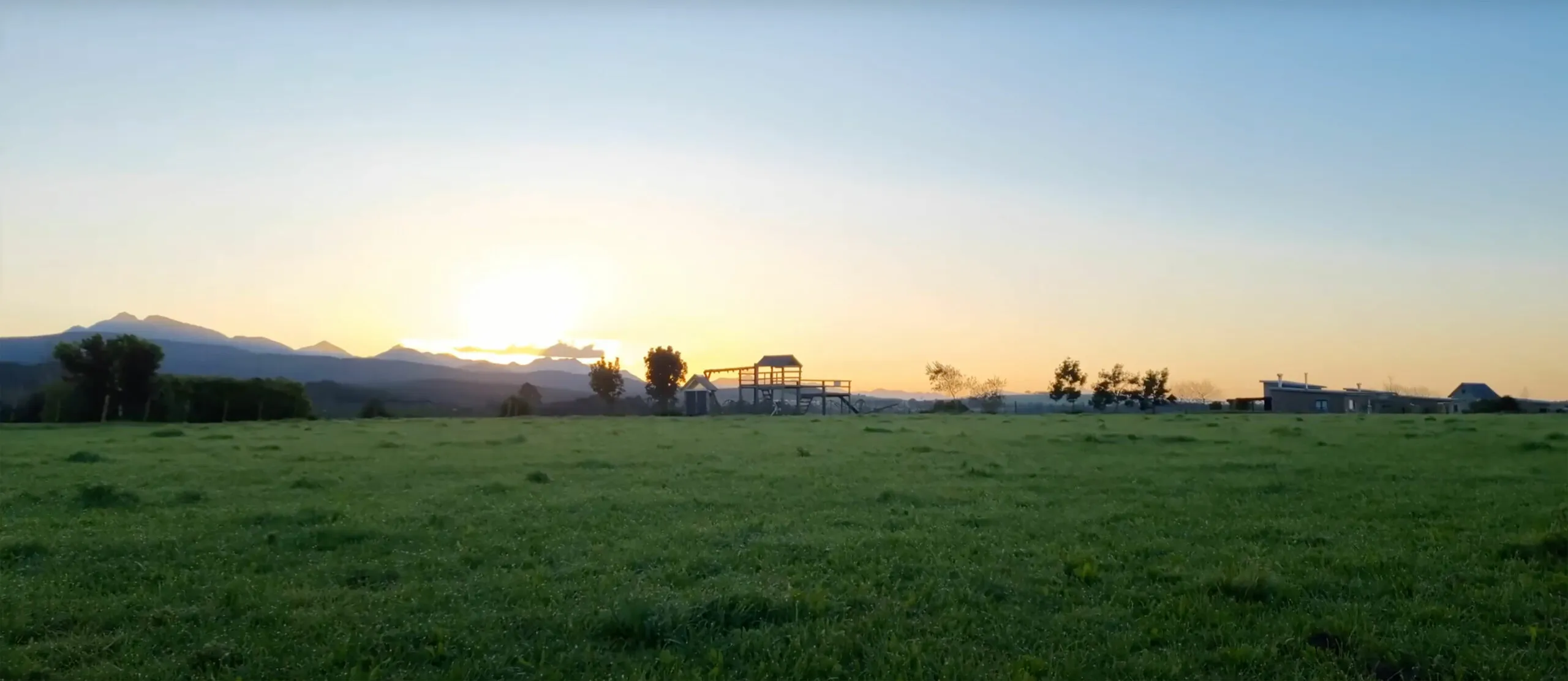 Sunset over a grassy field with distant mountains and a playground structure