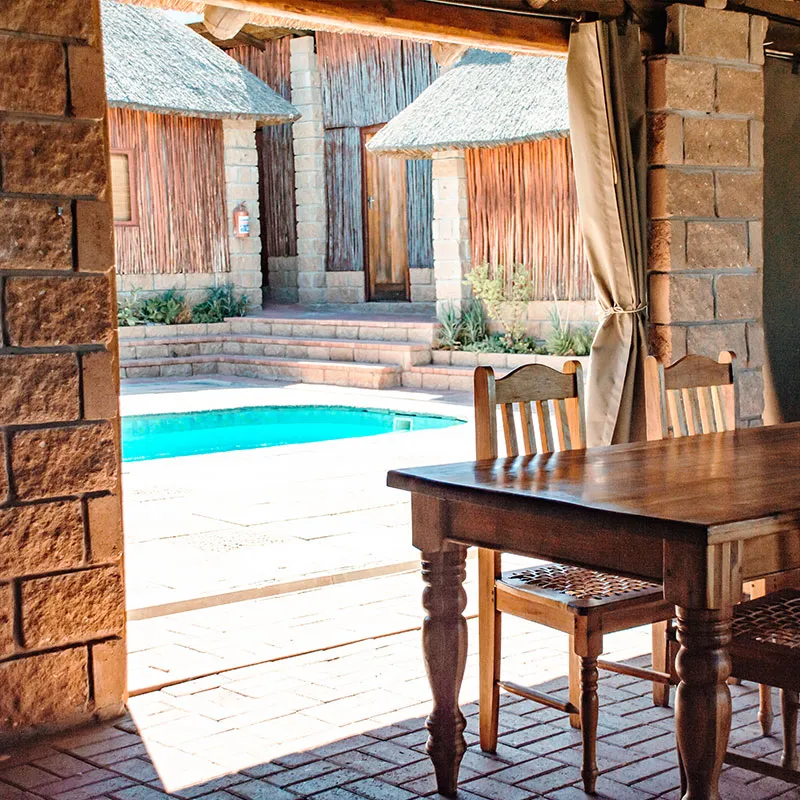 Wooden table and chairs by a pool with stone and thatched buildings in background