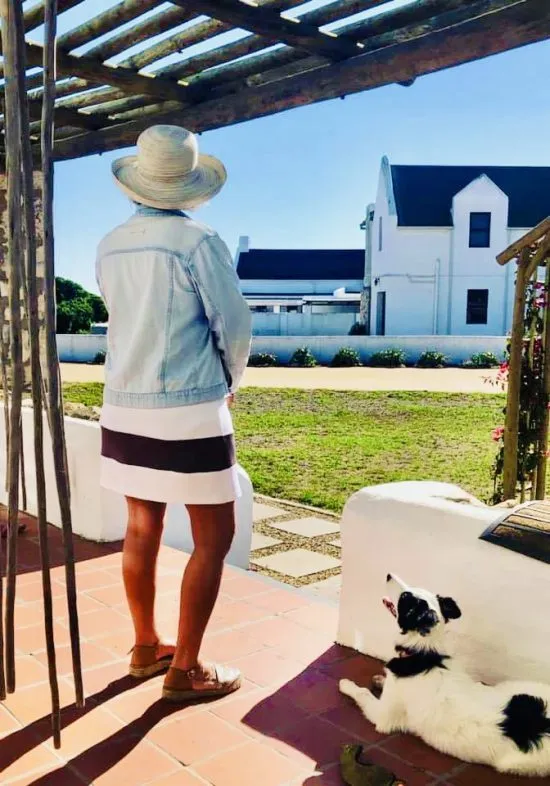 Woman in hat and dog on patio overlooking garden and white house