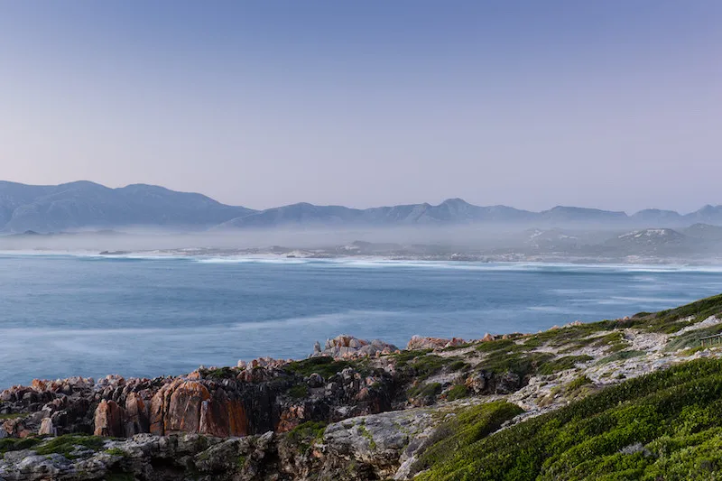 Coastal landscape with rocky cliffs green vegetation and distant mountains under a clear sky