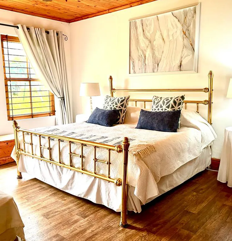 Brass bed with white bedding and pillows in a woodenfloored bedroom with large windows