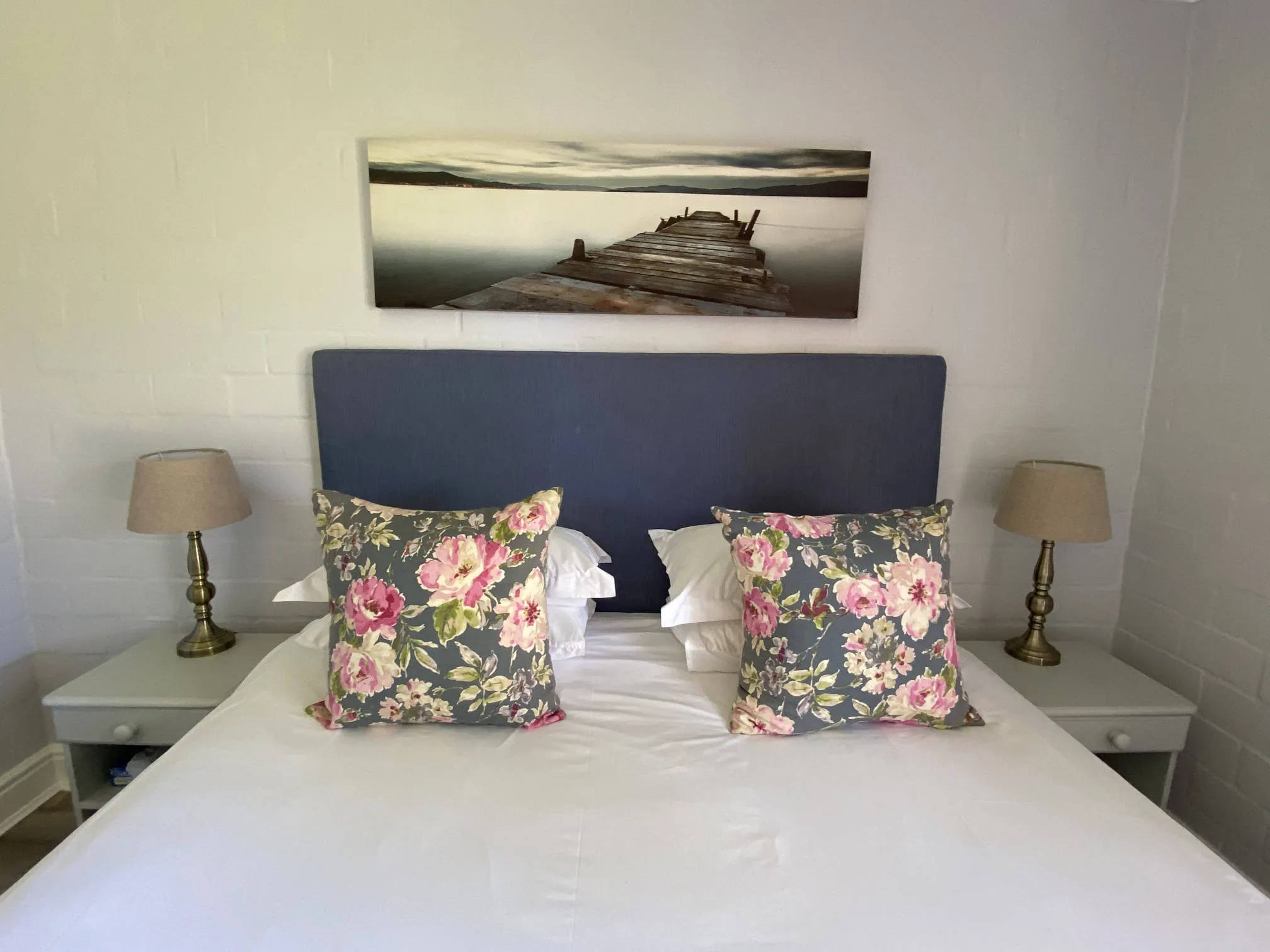 Bed with floral pillows two bedside tables with lamps and a wall painting