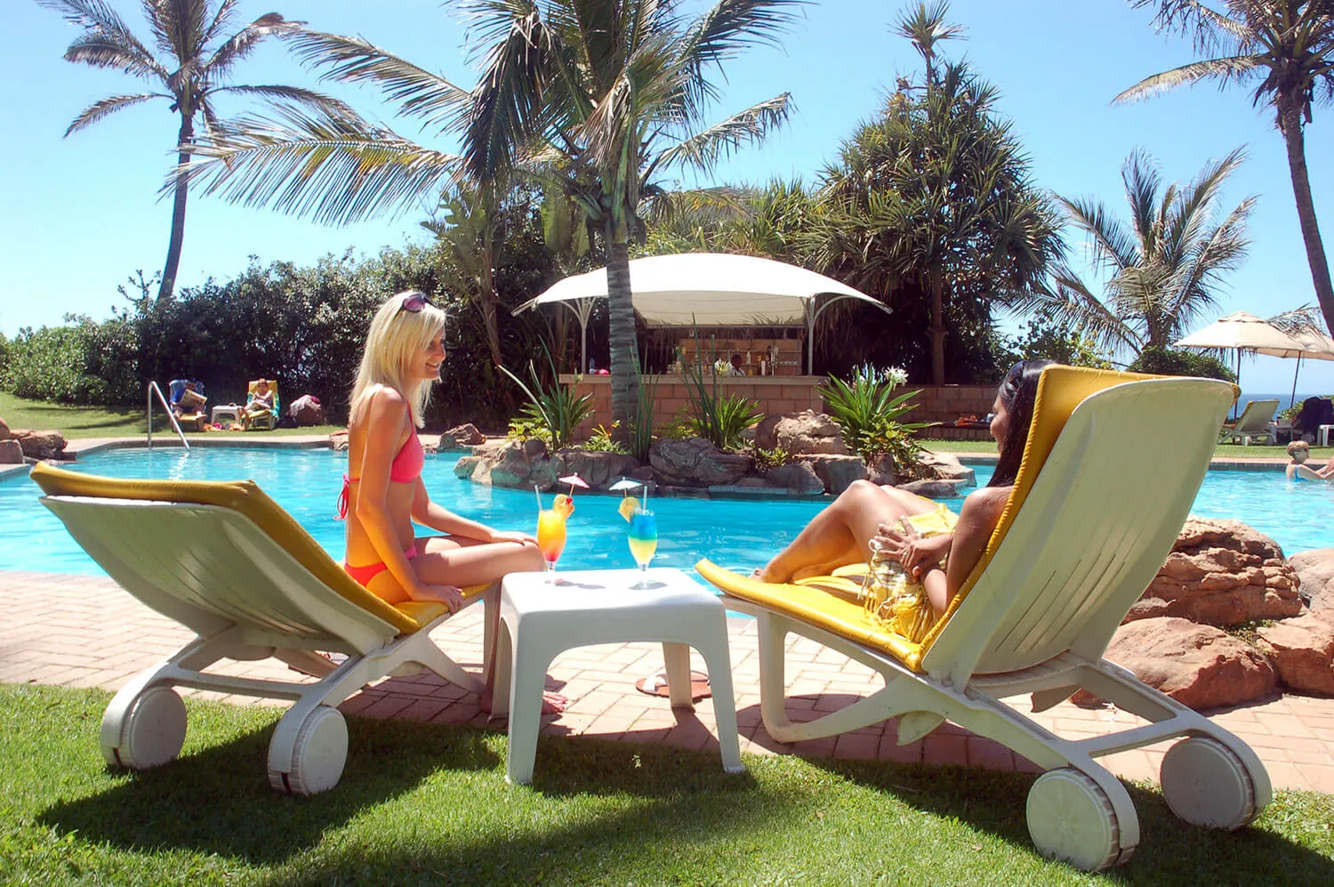 Two women in swimsuits relax on lounge chairs by a pool with drinks