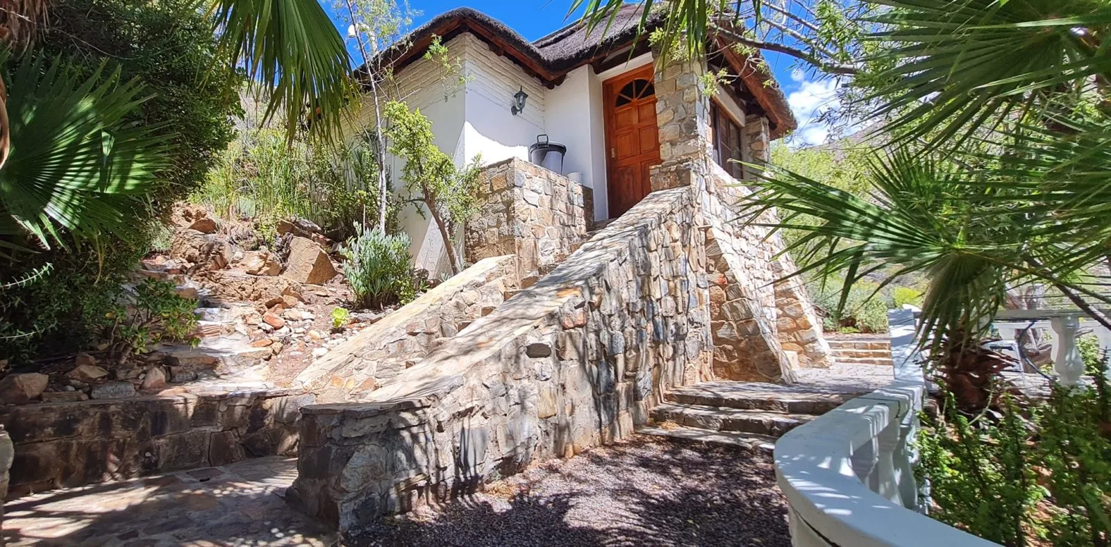 Stone and white house with wooden door surrounded by lush greenery and palm trees
