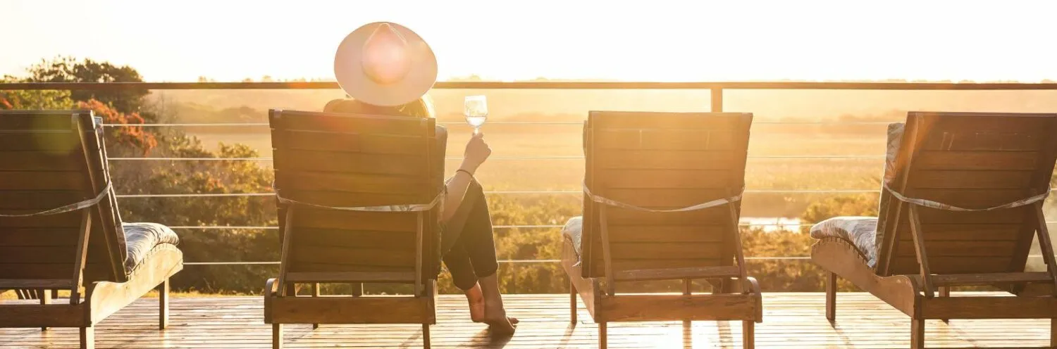 Person in hat sitting on lounge chair holding drink overlooking sunset landscape