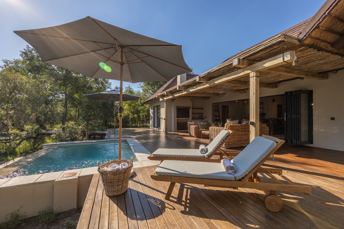 Wooden deck with lounge chairs, umbrella, and pool overlooking a garden and house.