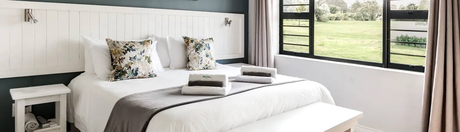 Bedroom with a neatly made bed floral pillows and large windows showing a green field