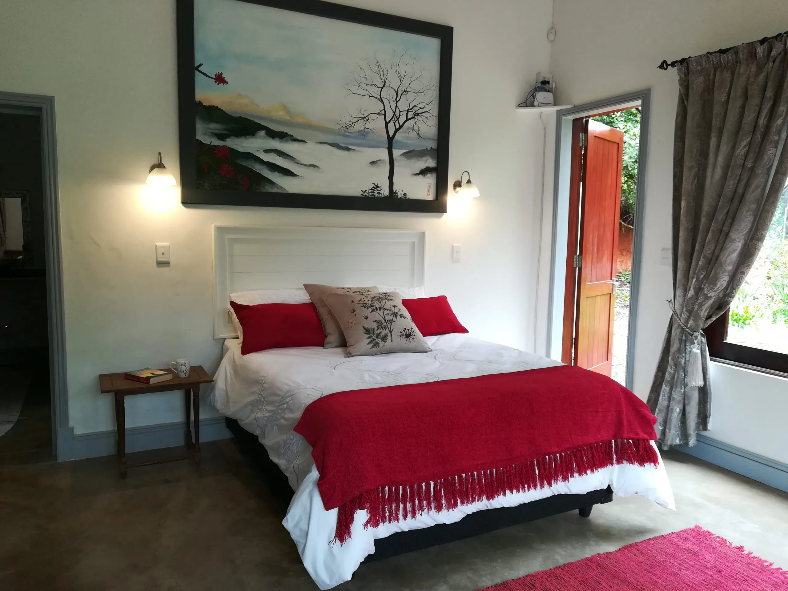 Bedroom with a bed red blanket large painting and open door to outside