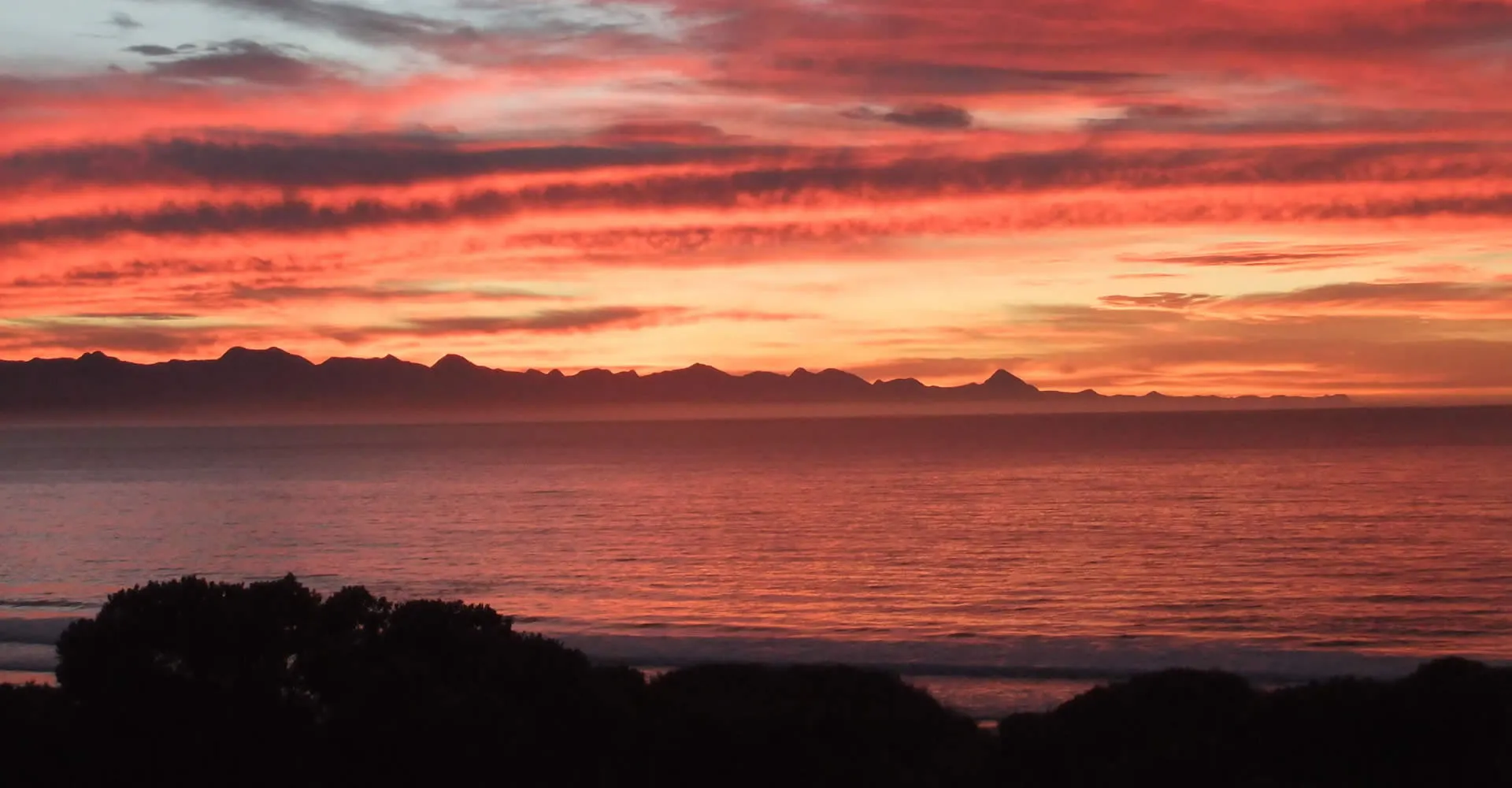 Sunset over the ocean with mountains in the distance and dark silhouettes of trees
