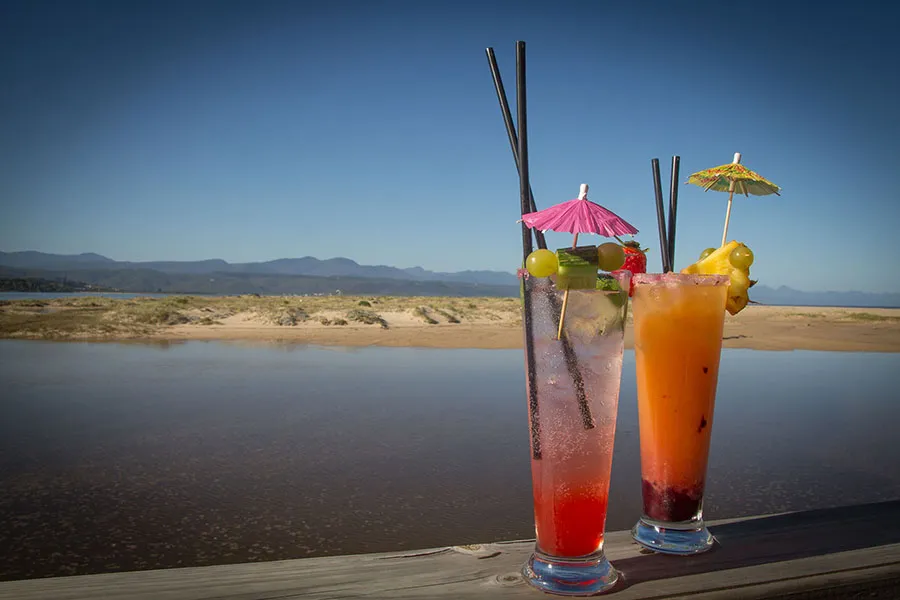 Two colorful cocktails with umbrellas by a lakeside with mountains in the background