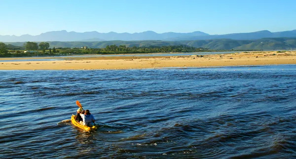 Two people kayaking on a river with a sandy shore and mountains in the background