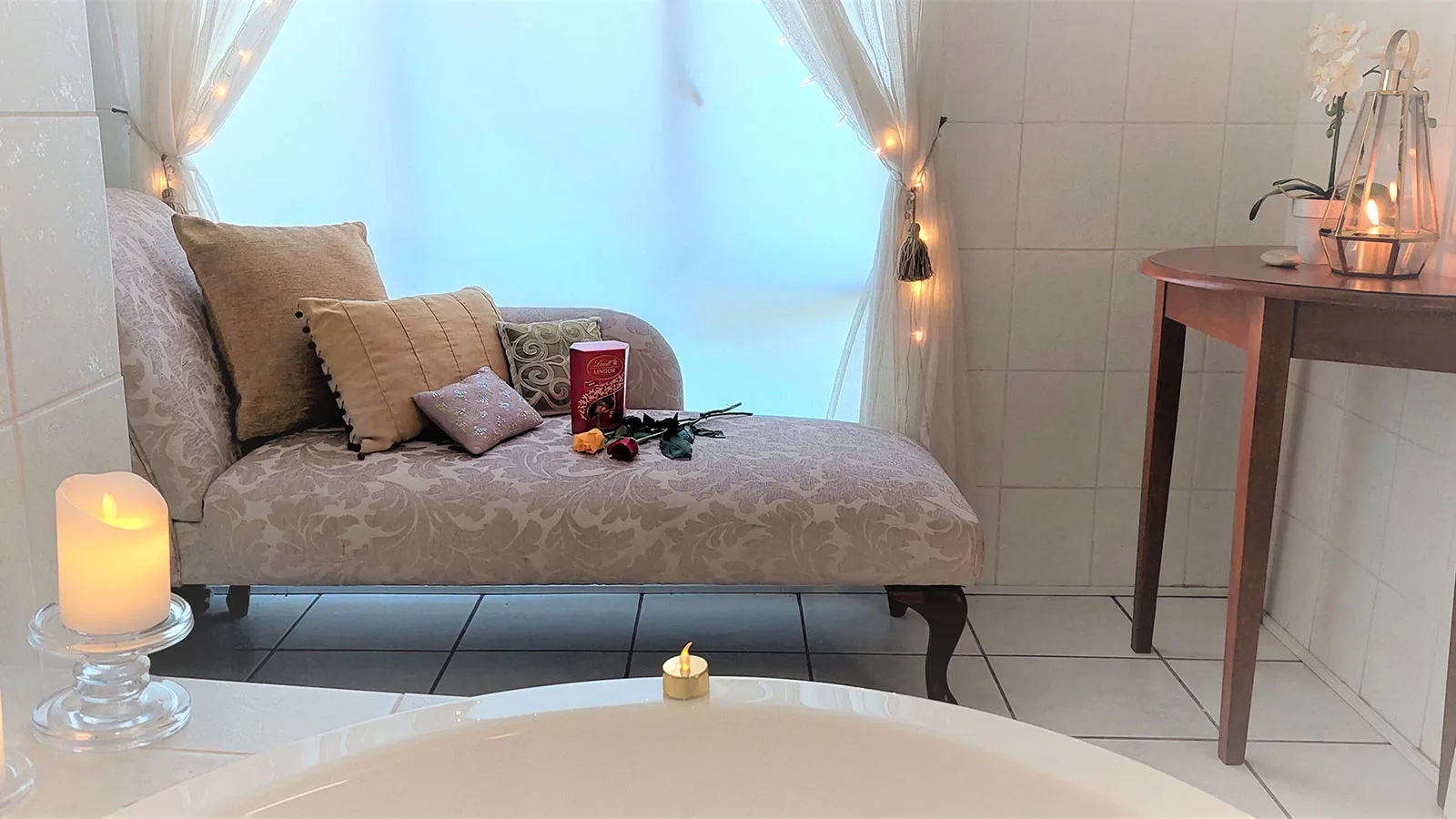 Chaise lounge with pillows book and candles by a window and bathtub