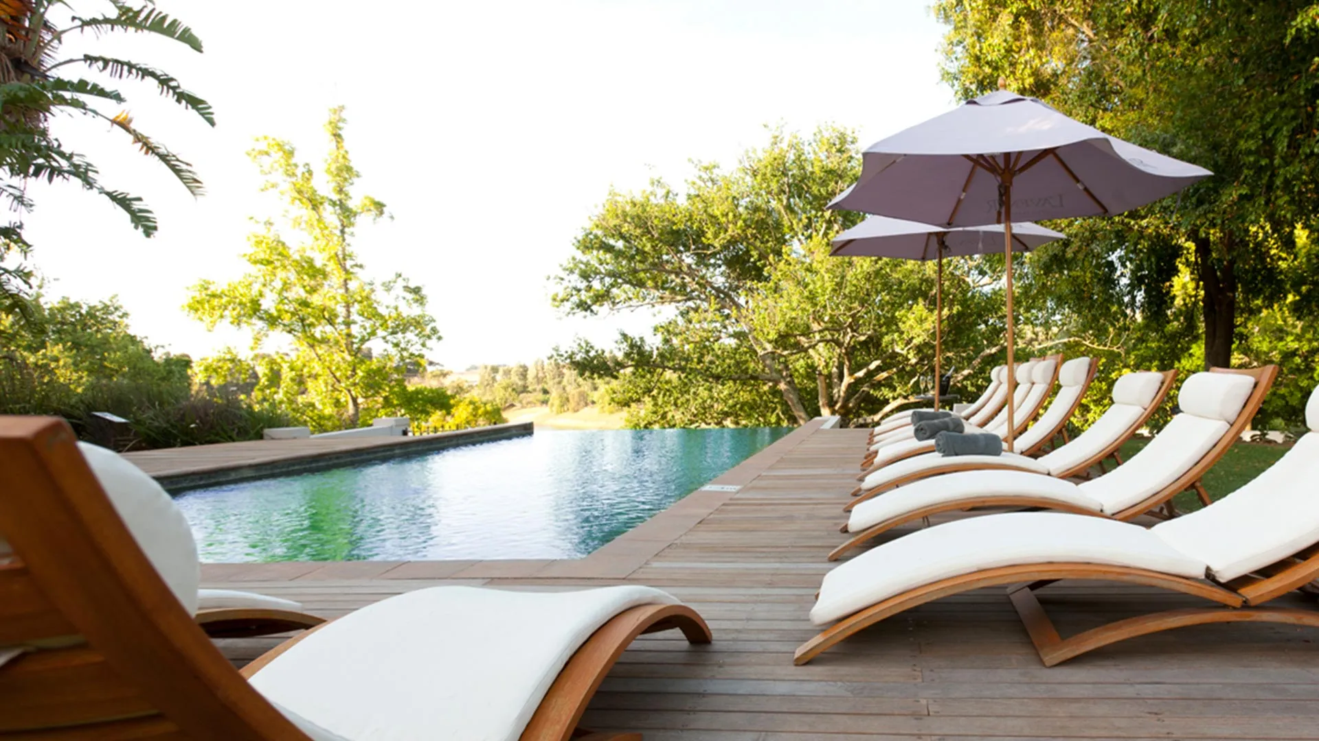 Row of lounge chairs by a pool with umbrellas in a lush garden