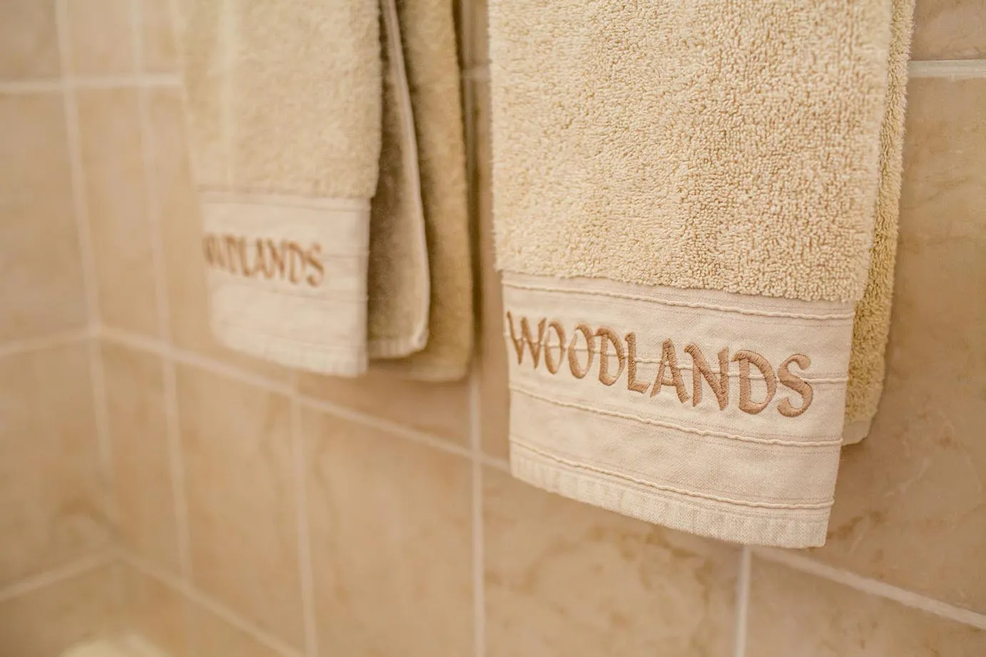 Two beige towels with WOODLANDS embroidered hanging on a tiled wall
