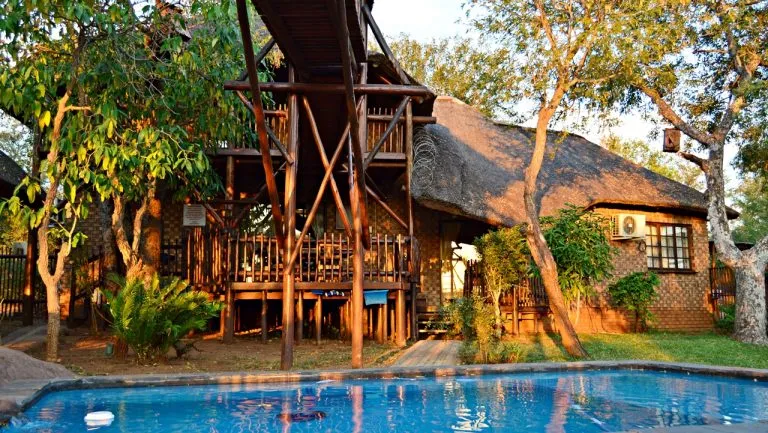 Wooden house with elevated walkway and pool surrounded by trees at sunset