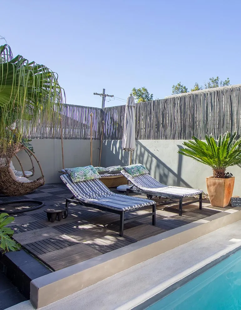 Two lounge chairs by a pool with plants and a wicker chair in a garden