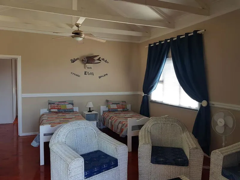Two single beds in a bedroom with wicker chairs ceiling fan and blue curtains
