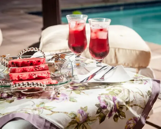 Two drinks and sliced watermelon on a floral tablecloth by a pool