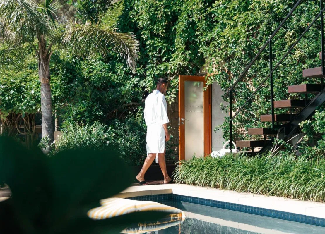 A person in a white robe walks towards a door near a pool and greenery