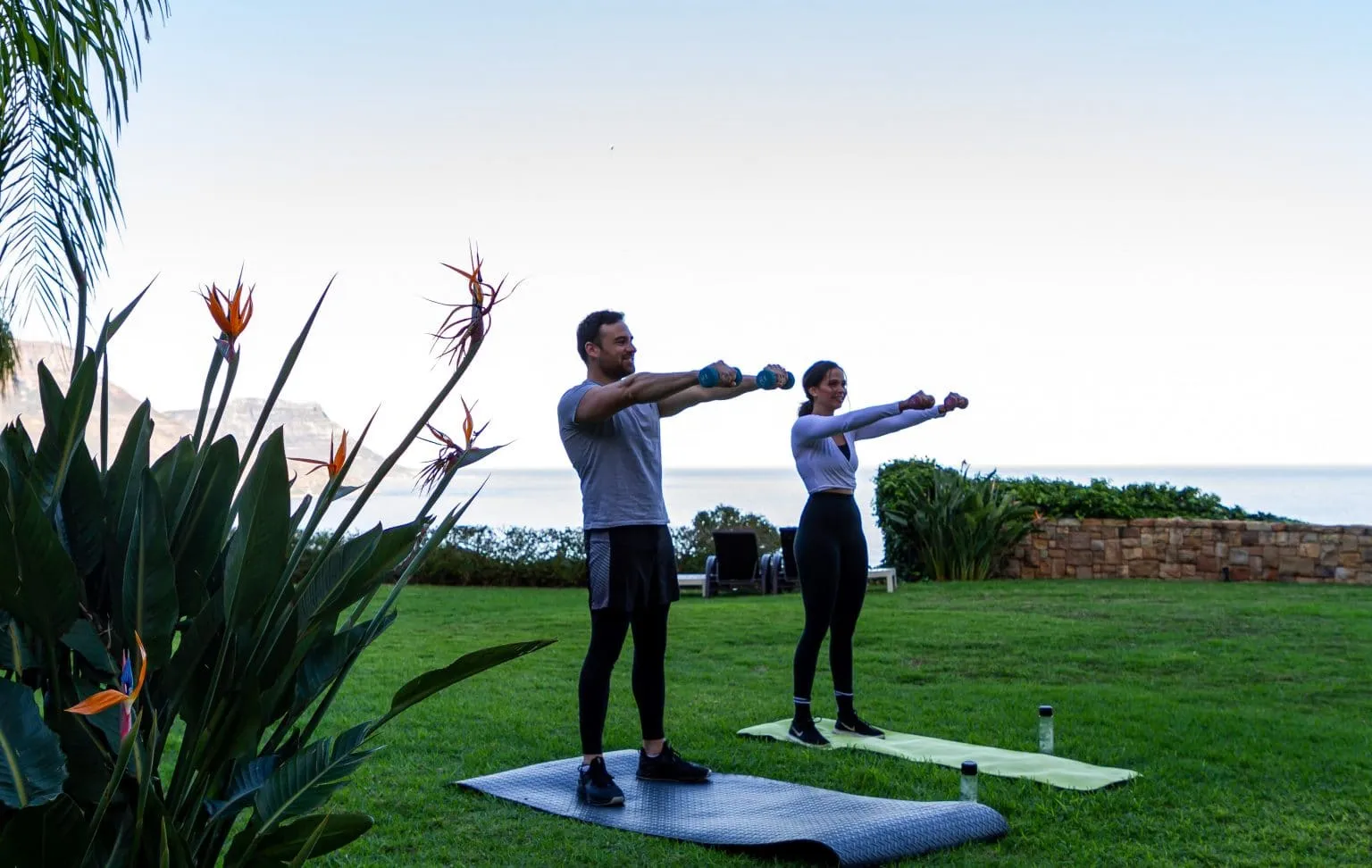 Two people exercising with dumbbells on yoga mats in a garden