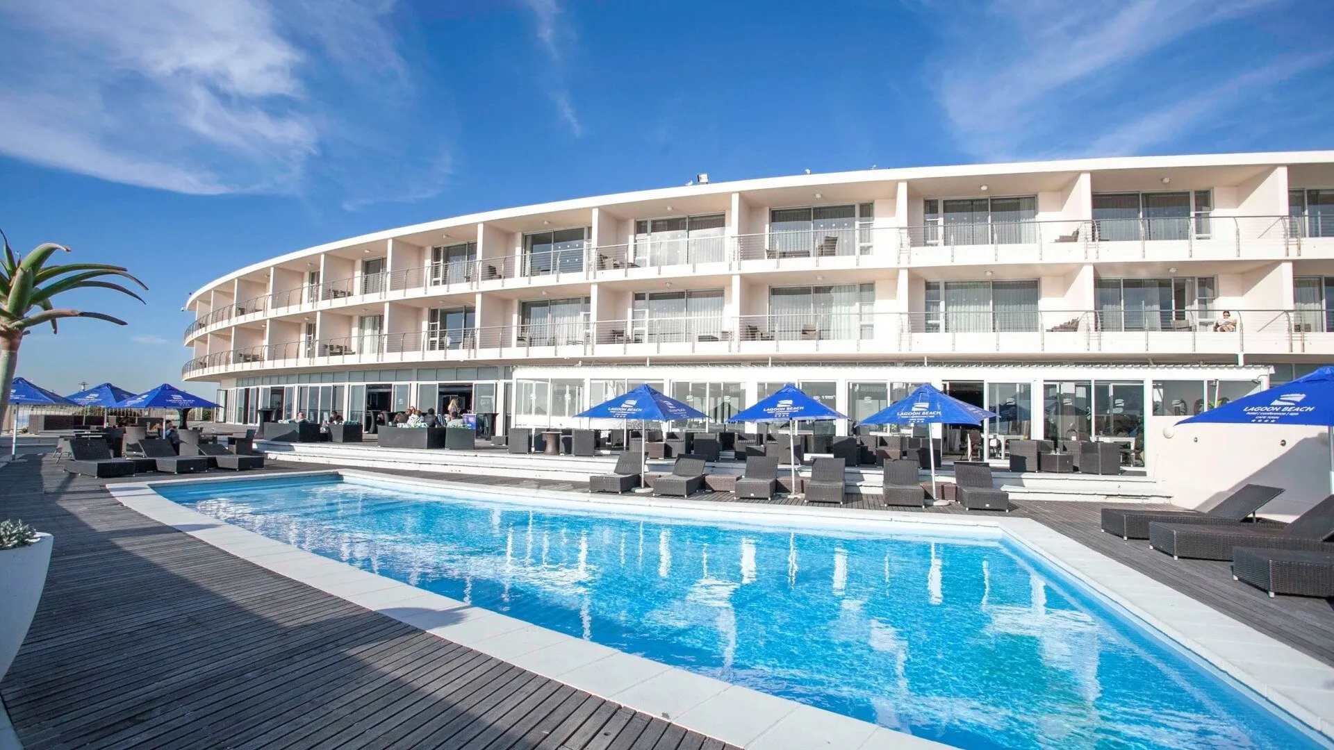 Modern hotel with pool lounge chairs and blue umbrellas under a clear sky