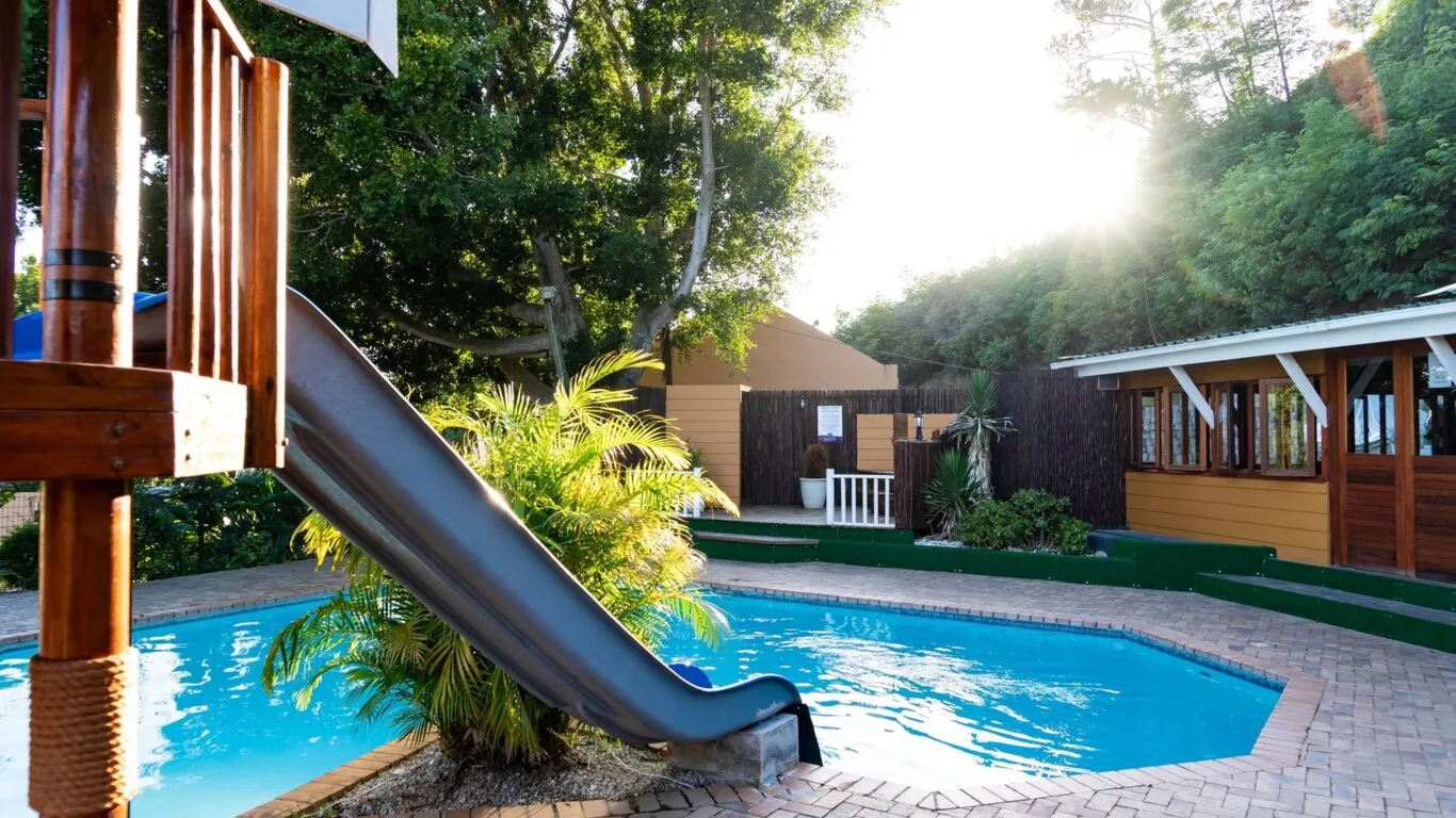 Wooden slide into a pool with lush greenery and buildings in the background