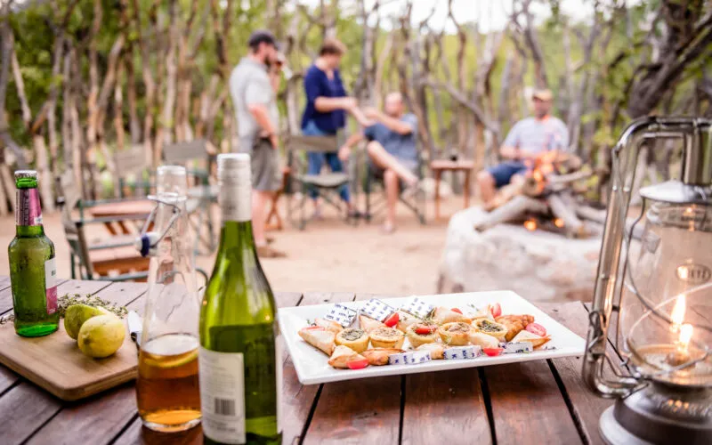 Outdoor gathering with food drinks and people around a campfire