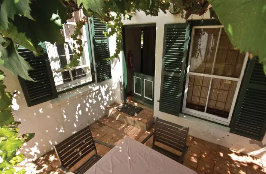 Patio with wooden chairs table and green shuttered windows under leafy shade