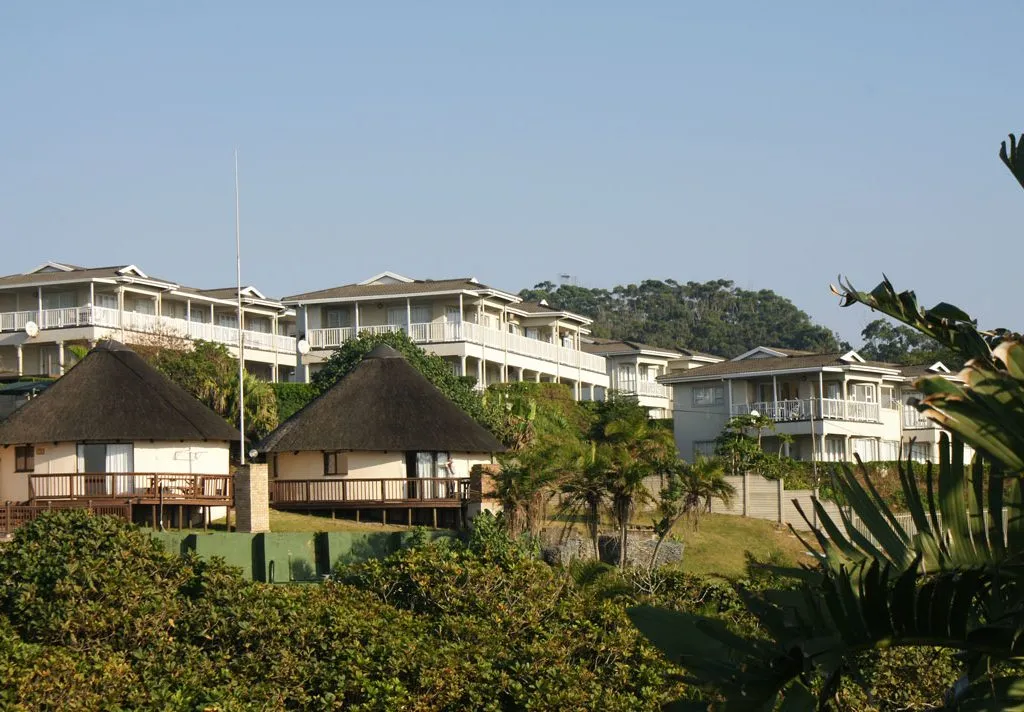Row of white resort buildings with thatchedroof huts in a lush green area