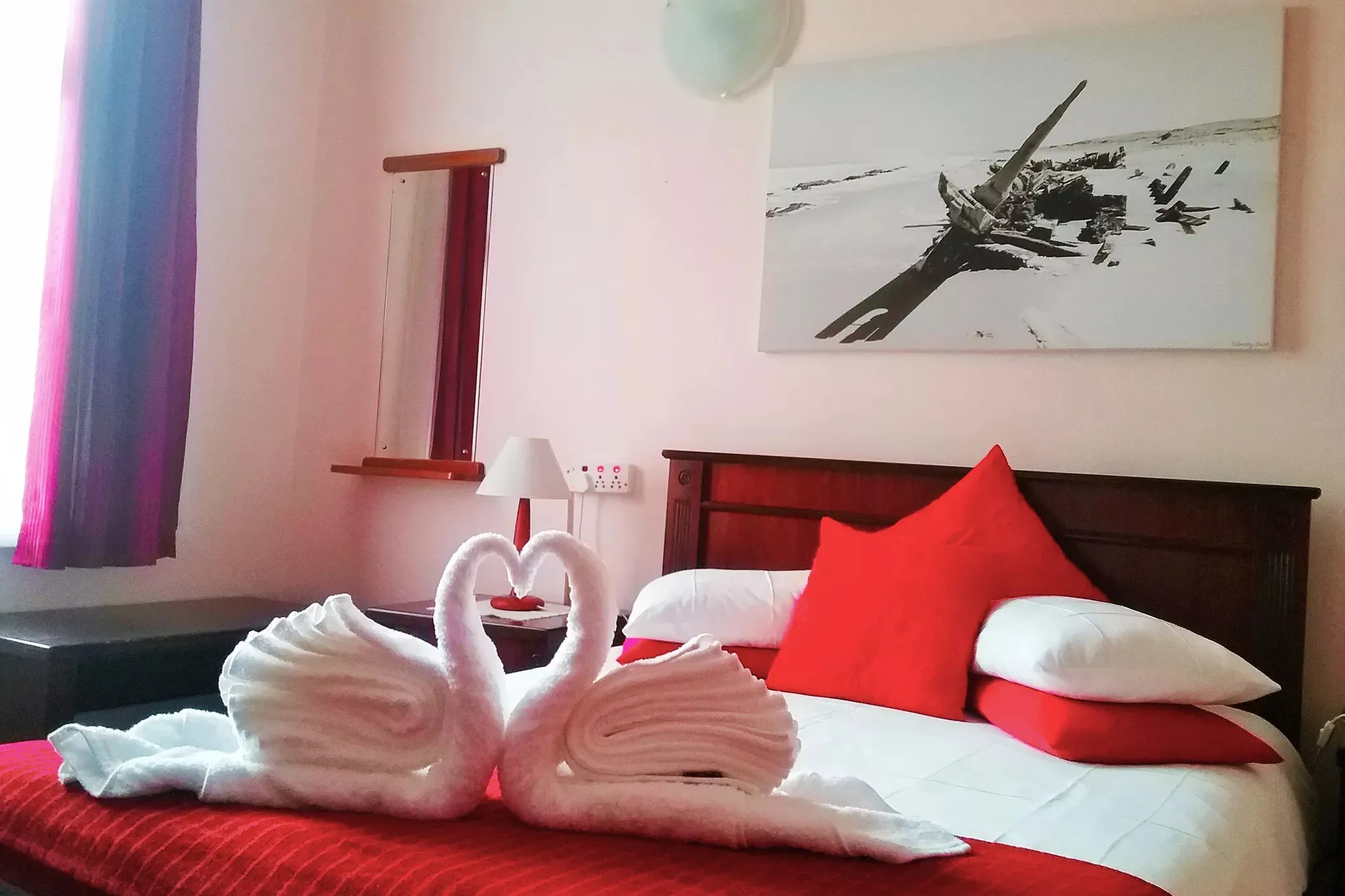 Bed with red pillows and towel swans framed picture above purple curtain