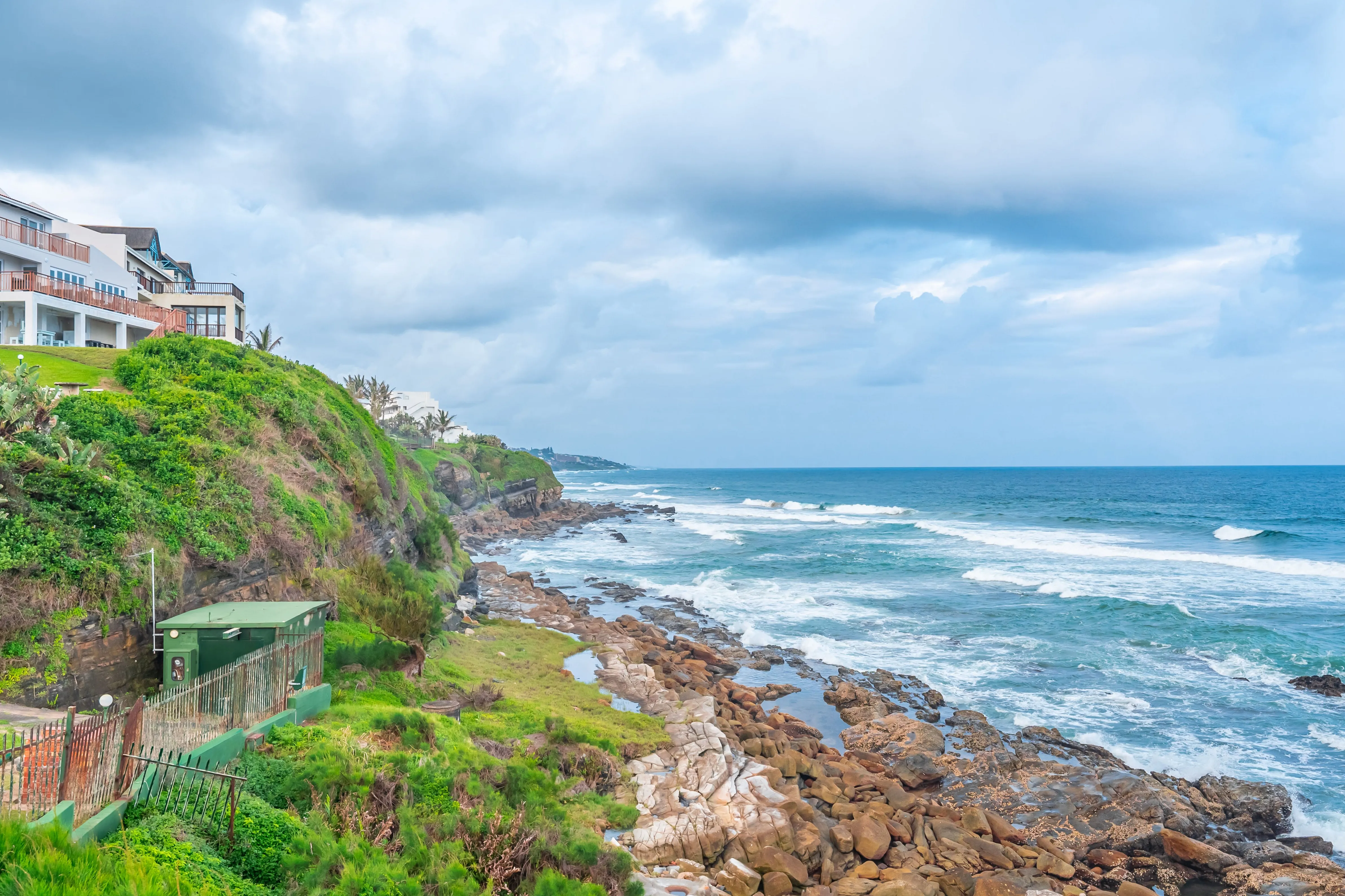Rocky coastline with green cliffs houses and ocean waves under a cloudy sky