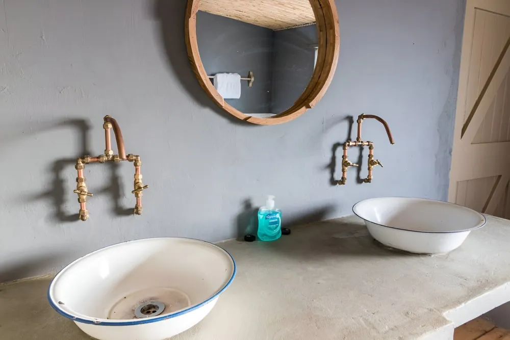 Two white sinks with copper faucets and a round mirror in a rustic bathroom